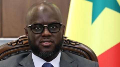 El Malick Ndiaye wears a suit and tie and sits in front of the Senegalese national flag.