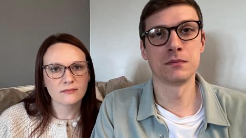 A woman with dark hair and glasses wearing a beige cardigan sits with a man with glasses on a sofa