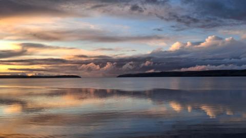 Calm coastal water reflecting a dramatic sunset sky, with soft orange and blue tones and layered clouds stretching across the horizon
