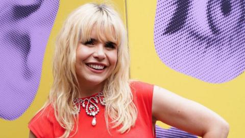 Paloma Faith smiles, she has bleach blonde wavy hair. She wears an orange tope and elaborate pearl and diamond necklace. She stands in front of a yellow and purple background.