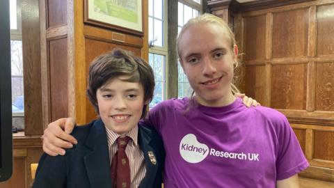 A teenage boy with dark brown hair wearing a maroon tie and red and white striped shirt and school blazer being hugged by an older boy with long, blond hair and wearing a purple t-shirt 