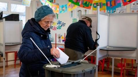 A woman with grey hair and a blue wooly hat places her ballot paper in a black box. She is clearly in a school classroom and there is a man in the background filling in his paper.