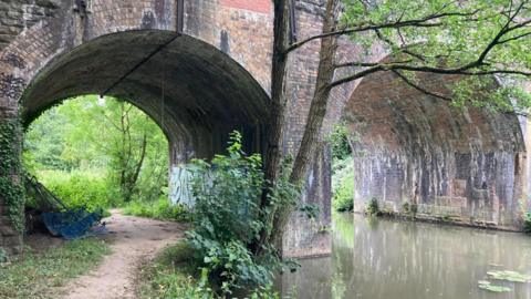 A section of path along the River Frome under the Network Rail Bridge. The footpath runs to the left of the river, with arches from the bridge above each.