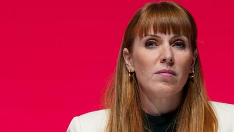 Angela Rayner who has long, ginger hair and is wearing a white suit and gold hooped earrings, looks to her right in front of a red background