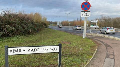 The A6, showing four lanes of traffic, with four vehicles on it. There is a large grass area, with a sign on it, with bushes in the distance, and several road signs. There are power lines overhead.