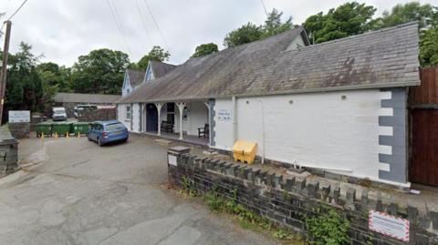 A white building with a sloped roof has a blue car parked outside.