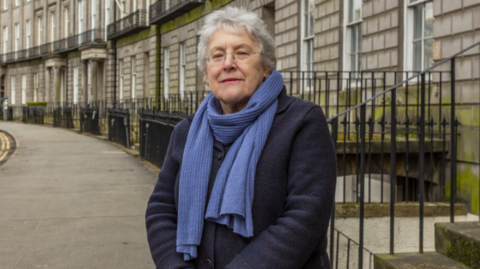 A woman with a light blue scarf stands in front of a row of Georgian houses in Edinburgh 