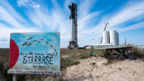 A sign that says "Live in our tomorrow today: Starbase, Boca Chica, TX" in front of a sand dune and a rocket launch facility in the distance.