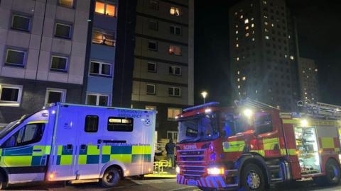 An ambulance and a fire engine sitting outside a block of flats. Lights are on in some of the building's windows.