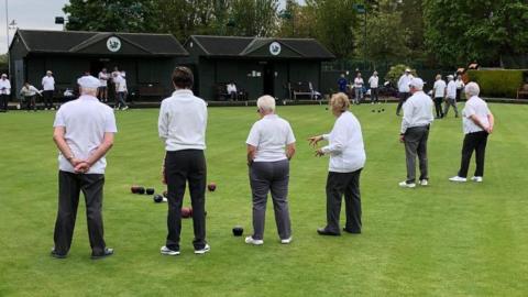 Members f the club playing bowls - wearing white shirts and black trousers, standing on the green grass.