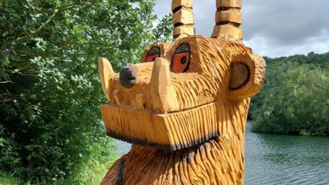 A wooden Gruffalo statue with trees and a river in the background. It had horns, orange eyes, and big teeth.