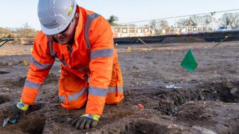 An archaeologist wearing fluorescent orange jackets and trousers, kneeling down, working on a site of bare earth with holes visible in the ground...