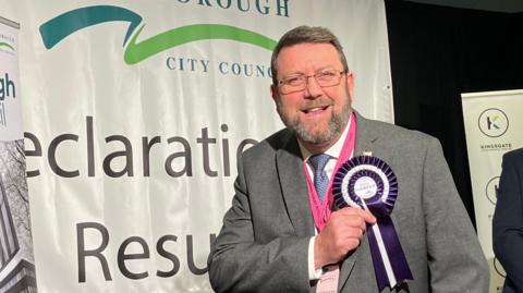 Chris Harper with a beard and glasses wearing a grey suit. He is touching his purple and white rosette with his name on it. A Peterborough City Council sign saying "Declaration Results" is in the background.  