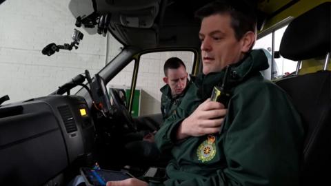 Inside the cab of a North East Ambulance Service vehicle. The paramedic in the passenger seat is speaking into a walkie talkie mounted on his shoulder. He is wearing a green paramedic uniform. A second paramedic on the driver side of the cab also wearing a green paramedic uniform is looking down to the back of the cab.