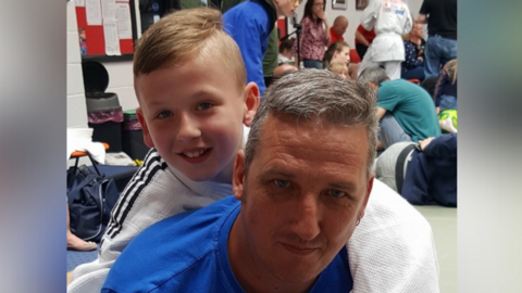 Simon Haycock wearing a blue T-shirt with his son Sam behind with his arm around him.