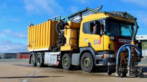 A lorry fitted with the specialist equipment used to carry out the work. The lorry is yellow and it has blasting equipment at the front, below the cab. 