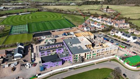 An aerial view of a school being constructed. There is purple panelling on one building and a crane lifting panelling on another. The school is surrounded by a new housing development and sports pitches.