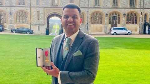 Clive Lawrence, wearing a suit and tie, holding up his OBE