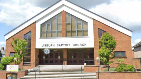 A triangle shaped brown bricked church with white overlayers and writing above the entrance that reads in capitals 'LISBURN BAPTIST CHURCH'. There are green trees at either side of the steps leading up to the entrance. 