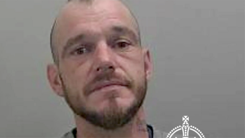 Mugshot of Luke Simon wearing a grey jumper. He has short hair and a stubble
