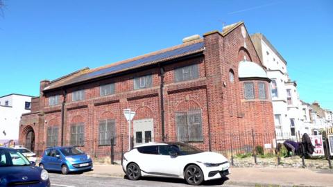An old brick building. Cars are parked outside. The sun is shining.