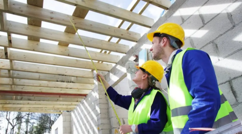 two men in high visibility vests and yellow helmets with a measuring tape, examining the wooden beams in the ceiling of a half-built house