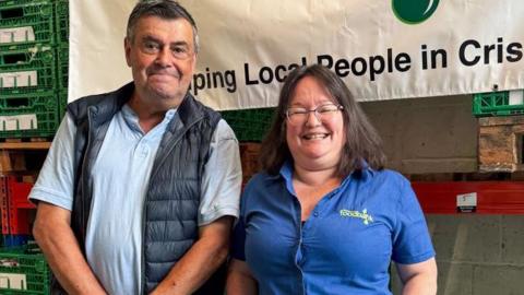 Mike and Maria smile at the camera while standing in a warehouse, part of a foodbank banner behind them. Mike is wearing a blue polo top and dark gilet while Maria wears a blue shirt with foodbank branding.