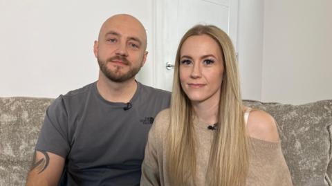 Tom and Becky Williams are sitting on a large, neutral coloured sofa. Tom has a tattoo on his arm and is wearing a dark grey top, he is bold and has a beard. Becky is wearing a neutral coloured top, she has blonde hair and together they are looking at the camera. Being them is the door to their living room.