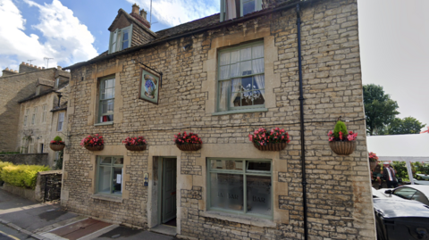 A two-storey Cotswold stone building, with green wooden sash windows on the upper floor. There are six baskets attached to the building with pink flowers in, and there is a swinging pub sign high up. 