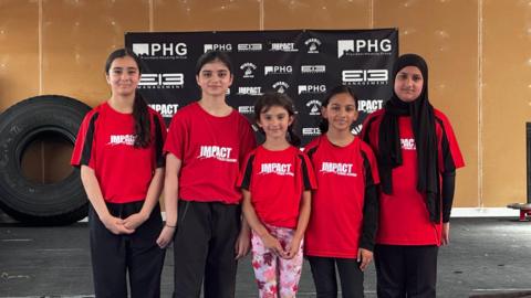 Five girls stand in a line smiling at the camera, wearing red t-shirts that say 'Impact Fitness Academy', in a room at the wrestling club