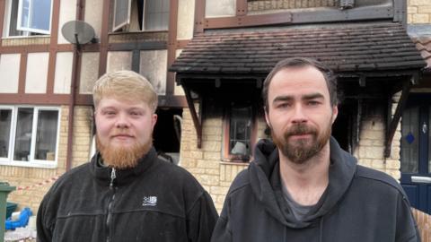 Liam and Harry stand in front of the burned remains of their home. the doors and windows are missing and there are black smoke stains up the yellow stone walls. They have beards and are wearing black jumpers