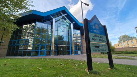 Basildon Crown Court is a large glass-fronted building with blue cladding and blue window trims. There is a sign in the foreground which says "Crown and County Courts", and there is a grass verge between the sign and the court building.