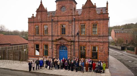 A large group of around 50 people stand together outside the front of a grand Victorian red brick building with ornate gables, a central tower and a bright blue arched doorway. The words "Free Library" are carved in stone above the entrance. The building is three storeys tall and flanked by other smaller buildings on what appears to be a recreated historic street.