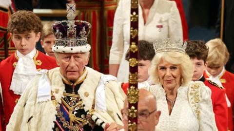 King Charles in his full royal regalia wearing a crown and white cloak over a military style suit and medals. The Queen is wearing a crown and dressed in right. She is to his left.