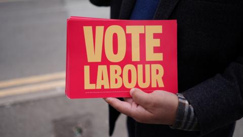 A close up picture of a persons arm holding a pile of vote labour flyers