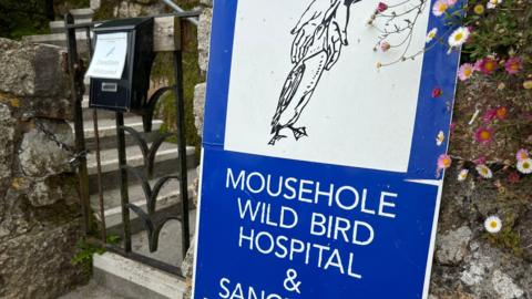 A blue sign with white writing that says MOUSEHOLE WILD BIRD HOSPITAL & SANCTUARY. Above the writing is a black and white line drawing of a bird with webbed feet and a long beak. The sign is on a stone wall next to a gate that is padlocked and has a donation box on it.