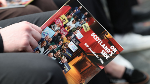 Close-up of a person holding a glossy booklet titled ‘Scotland on SNP Side – SNP Manifesto 2026,’ showing a collage of people standing on a stage, with the booklet resting on the person’s lap and other seated attendees blurred in the background