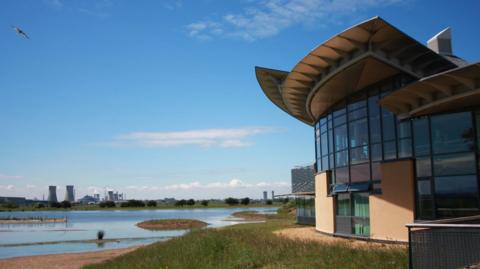 The glass fronted visitor centre at RSPB Saltholme. The building is surrounded by grass and looks out over water containing a number of small islands. Industrial cooling towers can been seen in the distance.