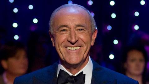 Headshot of Len Goodman. He is smiling and is wearing a dark suit and bowtie. In the background there are out-of-focus audience members and fairy lights