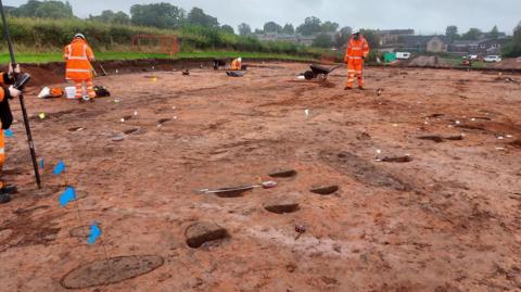 An archaeology site which has brown-reddish coloured earth. There are little blue flags and small parts which have been dug up. Two people wearing orange fluorescent overalls work on the site.