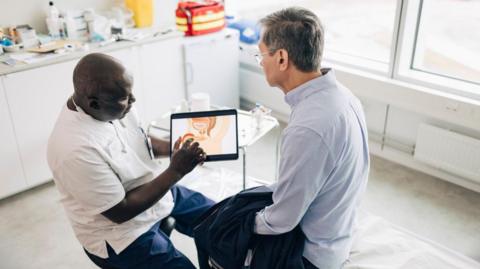 A bald man in a white top and navy trousers is holding and pointing at a digital tablet that has a male reproductive system on it. A man with grey hair and glasses and a blue shirt is sat next to him. Behind them is a medical room with equipment on a white table.