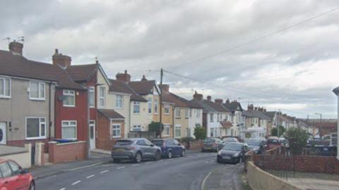 A residential street lined with a long row of attached and semi-detached houses on both sides of the road. The homes are predominantly two-storey properties with pitched tiled roofs, brick chimneys, and small front gardens or paved areas enclosed by low brick walls and fences.
In the foreground on the left, part of a red car is visible at the edge of the frame. Several other vehicles are parked along both sides of the street, including grey, blue, black, and silver cars. The road curves gently to the right as it continues into the distance.
The houses vary slightly in appearance but share a similar style. Most have cream, white, beige, or light yellow façades, while one prominent house near the front is painted a dark red colour with white trim. Many of the homes have bay windows, small porches, and brick details around the entrances.