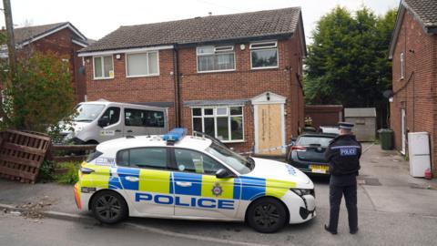 A police officer stands next to a police car in front of a semi-detached house which has been cordoned off with blue and white tape. The front door of the house been removed and replaced with a wooden board.