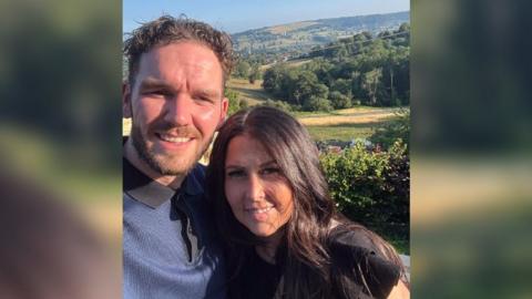 A couple, probably in their 30s, smile for a selfie in front of a stunning Cotswolds landscape, which is golden in the summer sun. The man is much taller than his partner, who he has his arm around, and has short, curly brown hair and is wearing a blue polo shirt with a black collar. She has long, wavy, dark brown hair, and a black top on. 