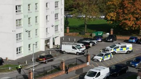 Police cars and other vehicles are parked outside a tower block. A police cordon is stretched across part of the scene and a small number of officers stand around it. 