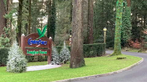 Wooden sign that reads Center Parcs Longleat Forest on a grass verge, with tall trees next to it. 