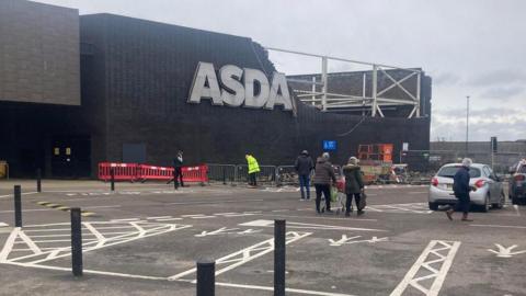 The outside of the Asda store in Hayle. A large section of the upper wall of the right-hand side of the modern-looking, two storey-high brown-bricked building is missing a large number of bricks, exposing a metal framework. The section is right beside the store's main "Asda" sign. Temporary fencing and red barriers have been put up around the damaged area. Several people are walking through the car park. A worker in a hi‑vis jacket is in the cordoned‑off section.