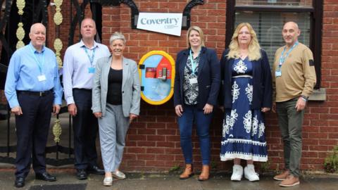 Three men and three women standing in front of a brick wall with a white sign which reads Coventry and below the sign is a yellow-framed box
