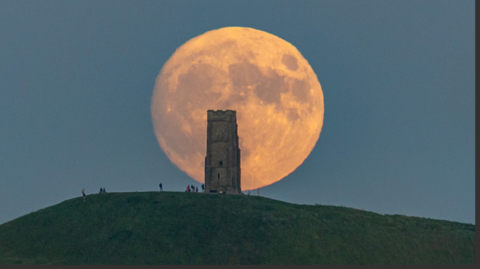 Glastonbury Tor at the top of a hill silhouetted by the huge strawberry-coloured full moon in a darkening blue sky.