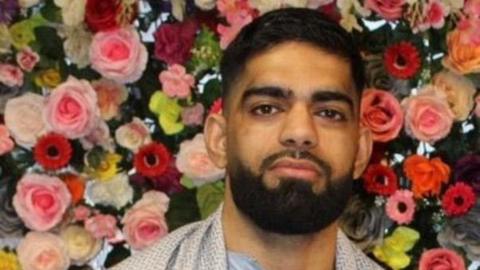 Luqman Ishfaq stands facing the camera against a backdrop of red and pink flowers